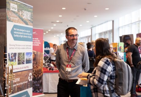 A delegate speaking to an exhibitor. Stands and other people can be seen in the background.