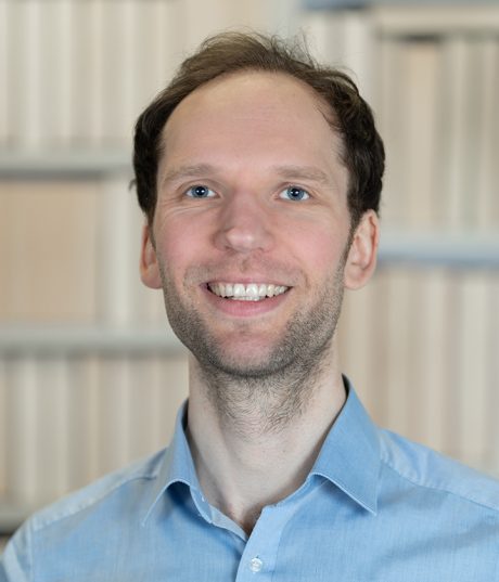 A photo of Linus Eusterbook, he is wearing a light blue shirt and standing in front of shelves. He is smiling at the camera. He has blue eyes and some facial hair.