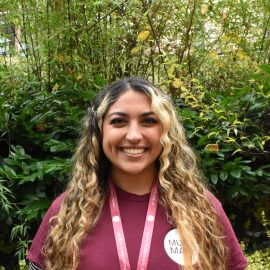 Aimee Christodoulou wearing a Music Mark t-shirt smiling at the camera, with foliage in the background.