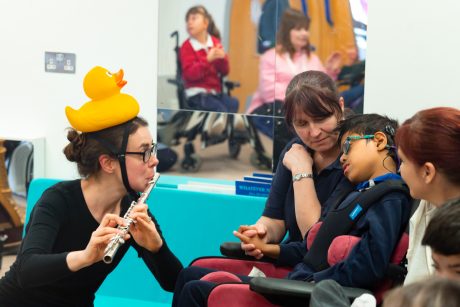 A musician wearing a rubber duck on their head plays the flute to a child sat with adults. A mirror in the background shows a reflection of other children in the room.