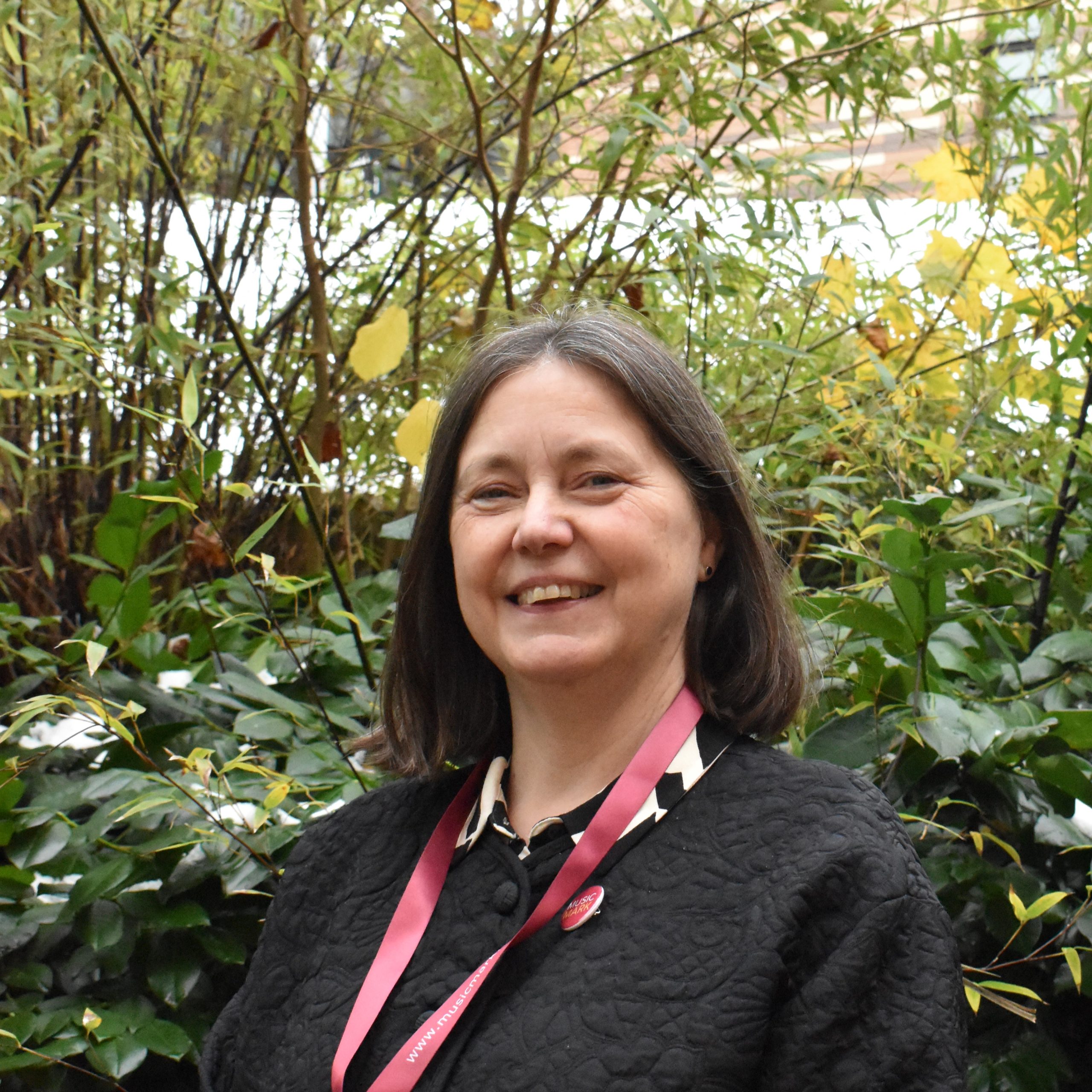 Bridget stood outside smiling at the camera. She has shoulder length brown hair, and is wearing a black jacket with a red lanyard.