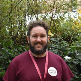 Chris Buglass wearing a Music Mark t-shirt smiling at the camera, with foliage in the background.