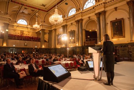 A speaker is stood on stage at a lecturn with their back to the camera. A grand hall filled with people sat at round tables listening to the speaker.