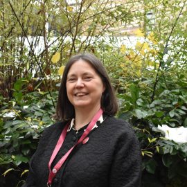 Bridget Whyte wearing a black quilted jacket and red lanyard smiling at the camera, with greenery in the background.