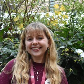 Emma Cragg wearing a Music Mark t-shirt smiling at the camera, with foliage in the background.