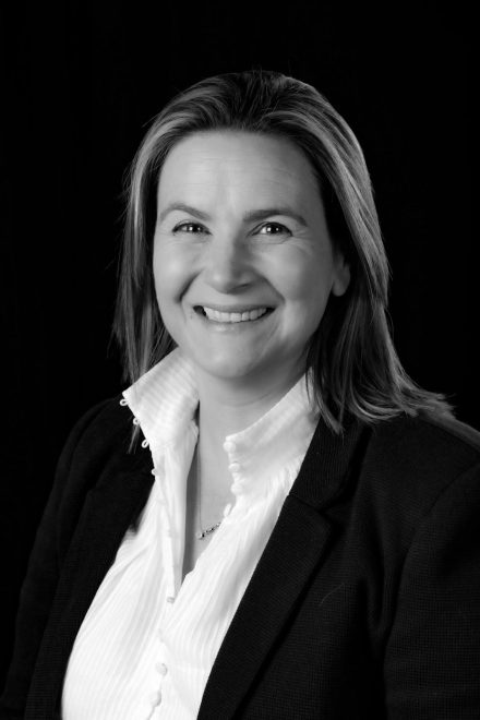 Black and white photo of Gemma Price. She is wearing a white shirt, a dark blazer, and is smiling at the camera.