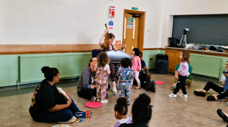 Adults are sat in a circle on the floor, with some children seated and other children stood around a woman holding a violin in the air