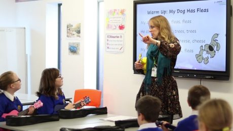 A teacher is stood at the front of the class holding a yellow ukulele. Several students are sat watching her, also holding ukuleles. A whiteboard can be seen behind the teacher reading 'Warm Up: My Dog Has Fleas'