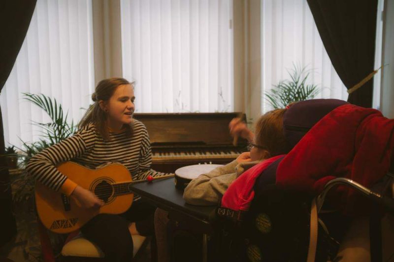 Beth Black is playing the guitar, they are sat in front of a piano. There is a person watching Beth play.