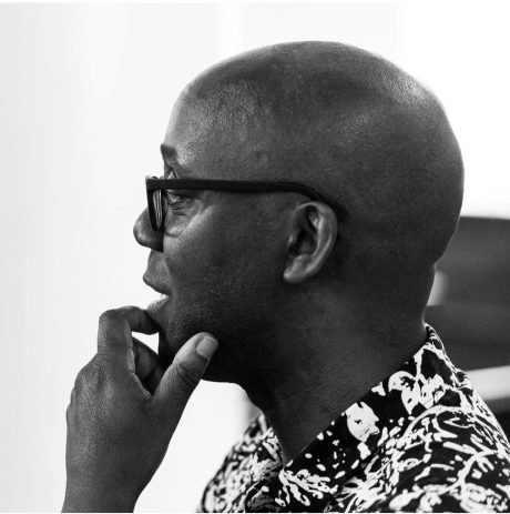 Robert Mitchell is dressed in a black and white shirt. He is looking away from the camera thoughtfully, holding his chin in his hand. He is wearing glasses.