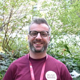 Jamie Munn wearing a Music Mark t-shirt and glasses smiling at the camera, with foliage in the background.