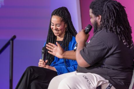 Dr Kadiatu Kanneh-Mason wearing a blue top holding a microphone, looking down smiling. In front of her is Rajae Wright, visible from the side, speaking with one arm up, the other arm holding a microphone