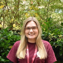 Katie Chappell wearing a Music Mark t-shirt and glasses smiling at the camera, with foliage in the background.