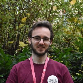 Matthew Crisp wearing a Music Mark t-shirt and glasses smiling at the camera, with foliage in the background.