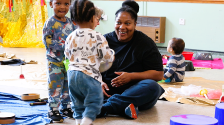 A woman sat on the ground smiling at a child facing away from the camera. Other children can be seen in the background. On the floor there are colourful pieces of fabric and various percussion instruments.