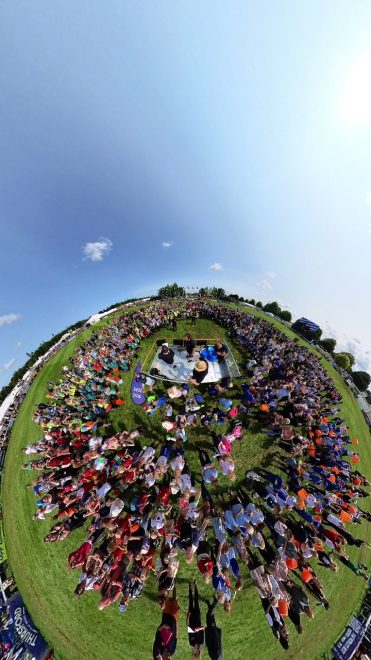Zoomed out image of a large group of people stood in a circle in a field, surrounding a small stage