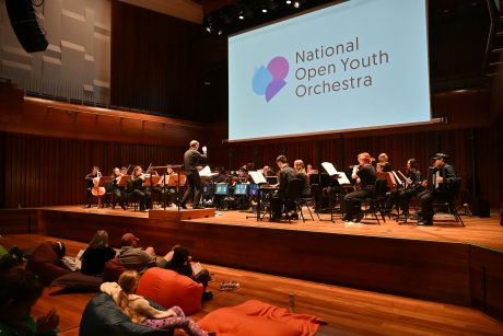 The National Open Youth Orchestra performing on stage. Some people can be seen sat on beanbags in the audience.