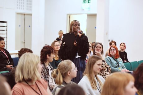 A young person stood holding a microphone and clipboard, surrounded by seated audience members.