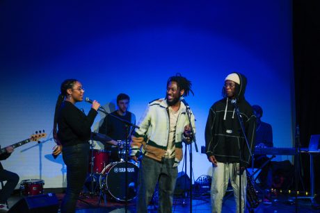 A group of musicians on stage - three singers stood at the front, a drumer behind, and obscured keyboard player and guitarist can be spotted. The background is blue.