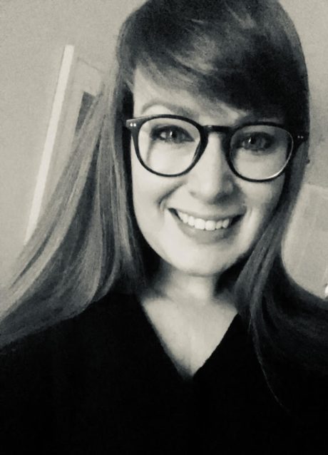 a black and white photo of Rachel, she is wearing a dark top, she has glasses, and long light hair. She has a fringe swept across the top of her glasses.