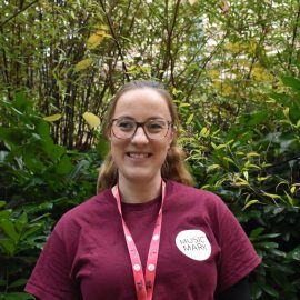 Rosie Lowe wearing a Music Mark t-shirt and glasses smiling at the camera, with foliage in the background.