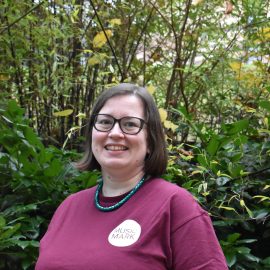 Sarah Whitfield wearing a Music Mark t-shirt, glasses and a turquoise necklace smiling at the camera, with foliage in the background.