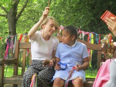 A young person sat on a bench outdoors next to a Sounds Wild volunteer, holding percussion instruments and craft items.