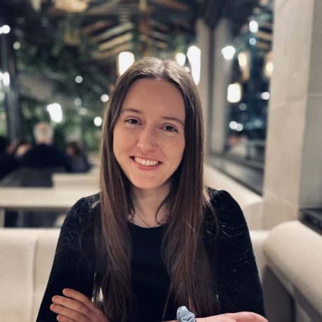 Tara Worrall-Hill smiling at the camera with her arms crossed on the table. She has long brown hair and is wearing a black long-sleeved top.