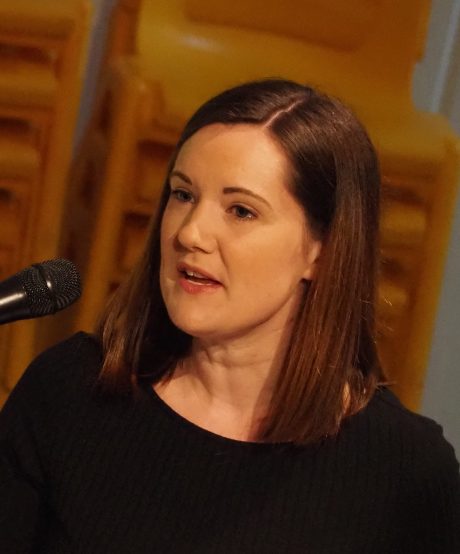 Tina Parsons speaking into a microphone, she has long brown hair and is wearing a black top.