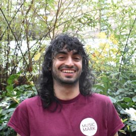 Yusef Sacoor wearing a Music Mark t-shirt smiling at the camera, with foliage in the background.