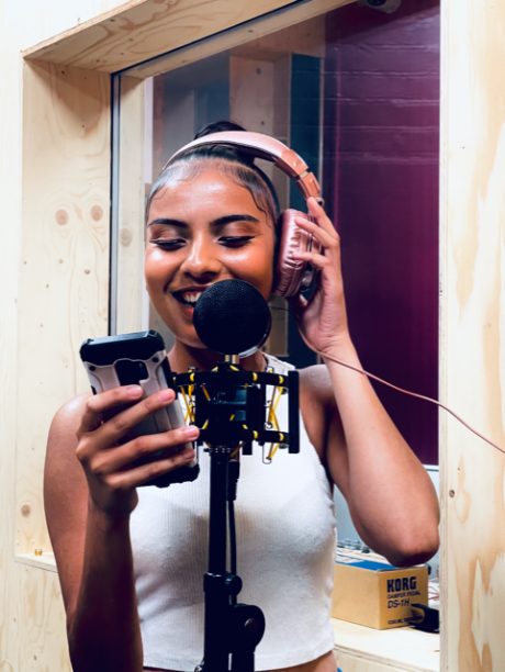 A young person in a recording booth, holding a phone in one hand and headphones to her ears with the other hand. She is smiling.