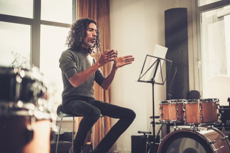 Person sat on a stool with hands in front looking at paper on a music stand. Drums are in the background.