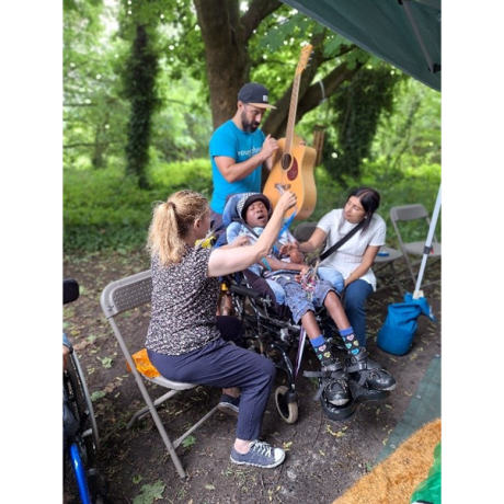 A young person in a wheelchair sat with three adults. One adult stood behind is holding a guitar up strumming the strings, whilst another holds another item up in the air.