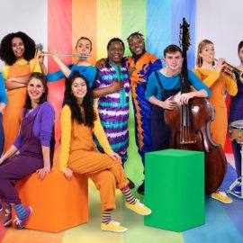 10 people wearing colourful clothes holding a variety of instruments in front of a rainbow background.