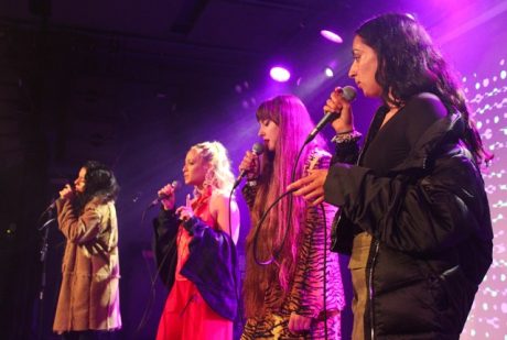 Four young people stood onstage singing, with a pinkish light.